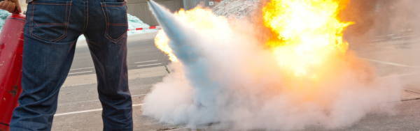 The image shows a person using a fire extinguisher to suppress a fire. The fire is producing bright flames and smoke, while the extinguisher releases a cloud of suppressant aimed at the fire. The person is standing to the left, partially visible, wearing jeans and holding the extinguisher.